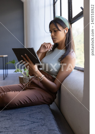 Asian woman sitting on bench near window in seating area, holding tablet with earbud, copy space Asian woman sitting on bench near window in seating area, holding tablet with earbud, copy space 140558922