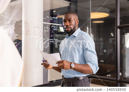 African american man drawing on glass whiteboard while holding papers and marker in office African american man drawing on glass whiteboard while holding papers and marker in office 140558930