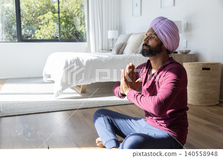 Mid adult indian man meditating pressing hands at chest in bedroom next to bed, window, copy space Mid adult indian man meditating pressing hands at chest in bedroom next to bed, window, copy space 140558948