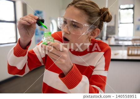 Female child in goggles and striped sweater, holding green jar, lifting dropper in science lab Female child in goggles and striped sweater, holding green jar, lifting dropper in science lab 140558951