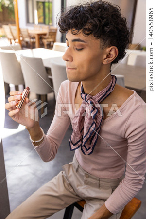 Male sitting on stool, holding lipstick tube near face wearing silk scarf 140558965