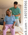Senior woman receiving shoulder massage from male caregiver on therapy table in living room 140558966