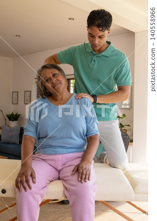 Senior woman receiving shoulder massage from male caregiver on therapy table in living room 140558966