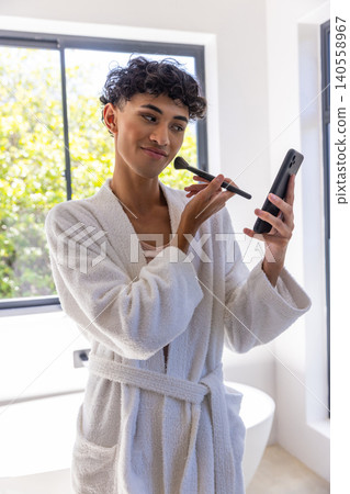 Asian man wearing bathrobe in bathroom holding smartphone, applying makeup brush by window 140558967