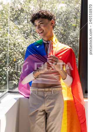Man leaning on windowsill wearing pink sweater and rainbow pride flag by window Man leaning on windowsill wearing pink sweater and rainbow pride flag by window 140558970