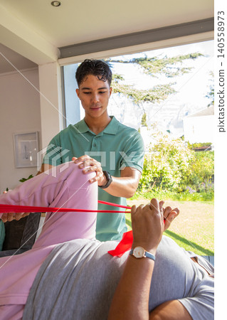 Asian physiotherapist assisting african american patient pulling red band in living room Asian physiotherapist assisting african american patient pulling red band in living room 140558973