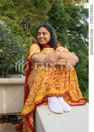 Indian woman sitting on stone ledge on terrace, hugging knees in yellow suit and gold bangles Indian woman sitting on stone ledge on terrace, hugging knees in yellow suit and gold bangles 140559002
