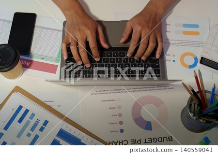 Hands typing on laptop with financial budget charts and documents on office desk Hands typing on laptop with financial budget charts and documents on office desk 140559041