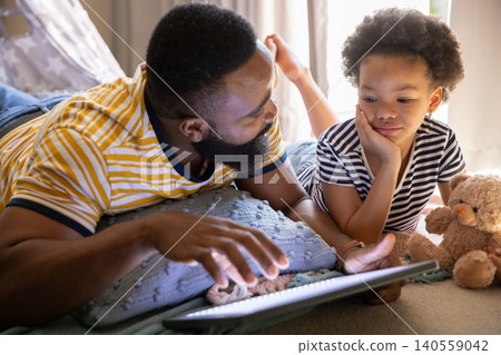 African American father and daughter lying on bed at home holding tablet alongside plush teddy bear African American father and daughter lying on bed at home holding tablet alongside plush teddy bear 140559042