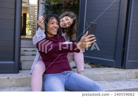 Diverse female friends taking selfie on concrete steps at entrance with smartphone keys, planter 140559062