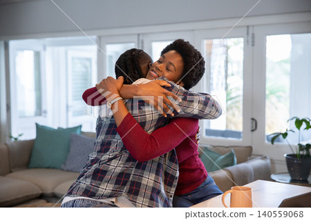 African american mother and daughter hugging, holding coffee mug beside table in living room African american mother and daughter hugging, holding coffee mug beside table in living room 140559068
