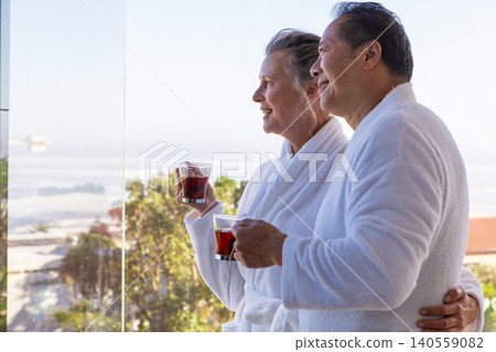 Diverse senior couple standing on balcony in robes holding mugs overlooking ocean, copy space 140559082