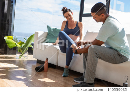 Diverse couple sitting on white sofa in living room tying teal athletic shoes by sea view Diverse couple sitting on white sofa in living room tying teal athletic shoes by sea view 140559085
