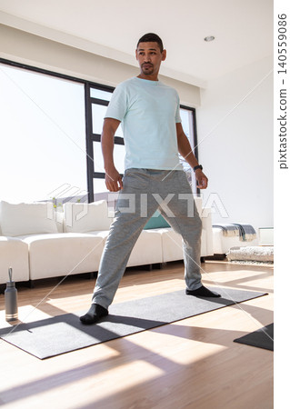 Man standing on yoga mat in living room stretching with water bottle and fitness tracker Man standing on yoga mat in living room stretching with water bottle and fitness tracker 140559086