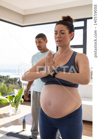 Pregnant woman practicing yoga with partner watching near water bottle, potted plant in living room Pregnant woman practicing yoga with partner watching near water bottle, potted plant in living room 140559088