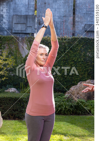Senior woman stretching arms overhead in backyard garden wearing rose top, smartwatch, leggings Senior woman stretching arms overhead in backyard garden wearing rose top, smartwatch, leggings 140559100
