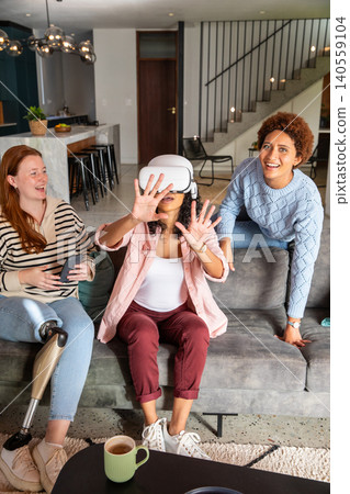 Diverse female friends playing virtual reality game on sofa in living room with controllers 140559104