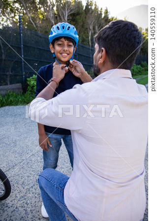 Asian father with son kneeling on paved path adjusting bright blue helmet strap on smiling boy Asian father with son kneeling on paved path adjusting bright blue helmet strap on smiling boy 140559110