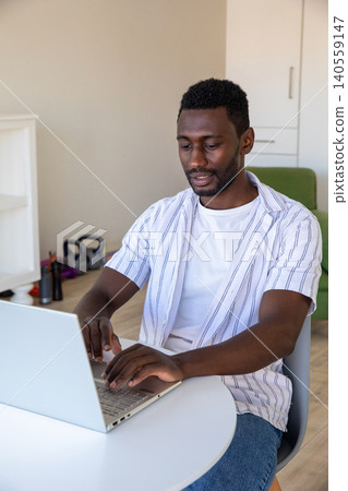 African american man working on laptop at white table in home office with kettlebells African american man working on laptop at white table in home office with kettlebells 140559147