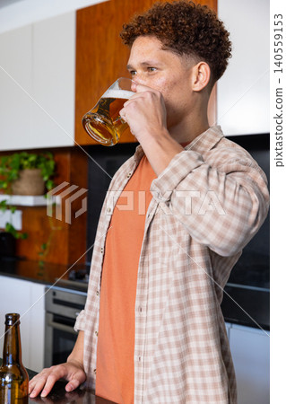 Man standing at kitchen stone countertop sipping beer from glass mug near bottle by plant Man standing at kitchen stone countertop sipping beer from glass mug near bottle by plant 140559153