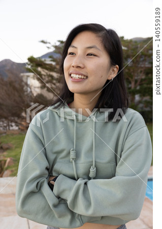 Asian woman standing with arms folded on backyard patio next to pool wearing hoodie Asian woman standing with arms folded on backyard patio next to pool wearing hoodie 140559189