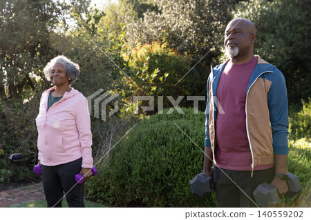 Senior couple in jackets exercising in garden lifting purple and black dumbbells Senior couple in jackets exercising in garden lifting purple and black dumbbells 140559202