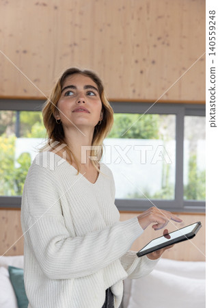 Woman standing near sofa in living room holding tablet and interacting with touchscreen at window Woman standing near sofa in living room holding tablet and interacting with touchscreen at window 140559248