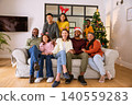 Diverse family gathering at home on sofa near christmas tree wearing santa hat, novelty glasses 140559283