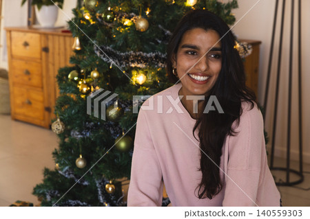 South Asian woman sitting next to decorated Christmas tree at home with ring lamp, copy space South Asian woman sitting next to decorated Christmas tree at home with ring lamp, copy space 140559303