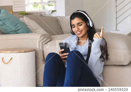 Indian woman wearing headphones at home, holding smartphone and leaning against couch near table 140559320