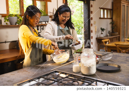 Diverse female friends wearing aprons whisking batter and adding cherries in home kitchen with bowl 140559321