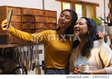 Diverse mother and daughter taking selfie in kitchen with smartphone mug and knife rack Diverse mother and daughter taking selfie in kitchen with smartphone mug and knife rack 140559322