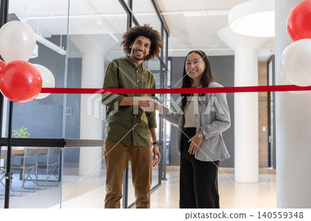 Diverse coworkers shaking hands across red ribbon in office lobby with balloons, glass partition Diverse coworkers shaking hands across red ribbon in office lobby with balloons, glass partition 140559348