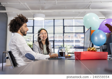 Diverse coworkers sitting at desk in open-plan office celebrating gift box, balloons by computer Diverse coworkers sitting at desk in open-plan office celebrating gift box, balloons by computer 140559354