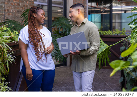 Diverse coworkers discussing project while standing in corridor with laptop near railing, plants Diverse coworkers discussing project while standing in corridor with laptop near railing, plants 140559377