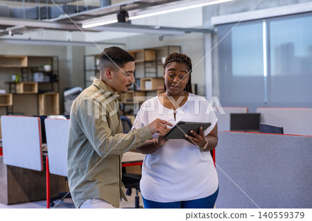 Diverse coworkers discussing details on tablet at desks in office with gray cubicle partitions Diverse coworkers discussing details on tablet at desks in office with gray cubicle partitions 140559379