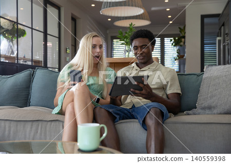 Diverse couple sitting on couch reacting to smartphone while man holding tablet in living room Diverse couple sitting on couch reacting to smartphone while man holding tablet in living room 140559398