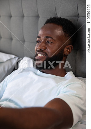 African American man reclining on bed with grey tufted headboard and white pillows smiling African American man reclining on bed with grey tufted headboard and white pillows smiling 140559403