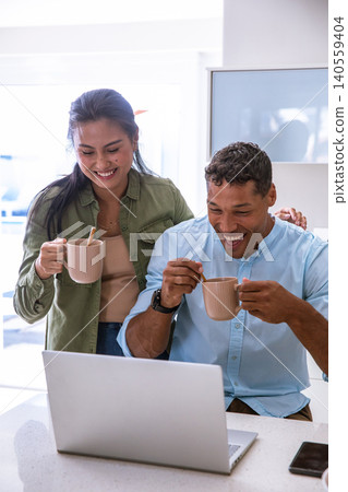Diverse couple laughing while standing at home kitchen counter with laptop, mugs with spoons Diverse couple laughing while standing at home kitchen counter with laptop, mugs with spoons 140559404