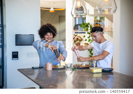Two male friends seasoning avocado toast and eggs with pepper grinders in open-plan home kitchen 140559417