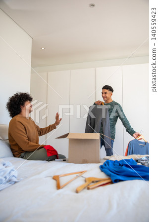 Diverse male friends sorting clothes into open cardboard box on bed in bedroom with wooden hangers Diverse male friends sorting clothes into open cardboard box on bed in bedroom with wooden hangers 140559423