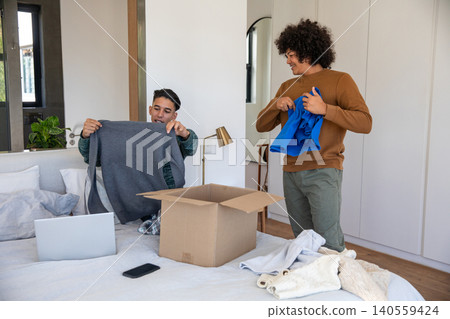 Diverse male friends unpacking gray sweater and blue shirt from box on bed in bedroom Diverse male friends unpacking gray sweater and blue shirt from box on bed in bedroom 140559424