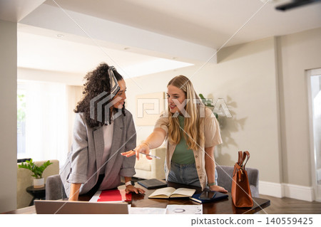 Diverse female coworkers leaning over table in home office examining laptop, documents Diverse female coworkers leaning over table in home office examining laptop, documents 140559425