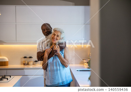 Diverse senior couple hugging in modern kitchen featuring glass jars bread box and gas cooktop Diverse senior couple hugging in modern kitchen featuring glass jars bread box and gas cooktop 140559436