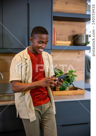 African American man leaning against blue kitchen cabinets, using smartphone beside tray with plant African American man leaning against blue kitchen cabinets, using smartphone beside tray with plant 140559438