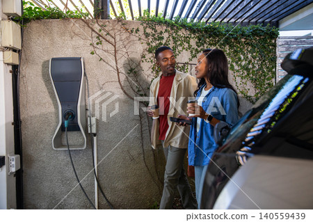 African American couple walking holding coffee cups checking smartphone by EV charger in carport African American couple walking holding coffee cups checking smartphone by EV charger in carport 140559439
