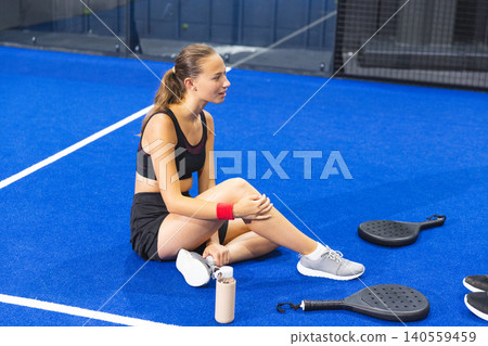Female athlete sitting on padel court in gear examining knee with rackets, water bottle Female athlete sitting on padel court in gear examining knee with rackets, water bottle 140559459