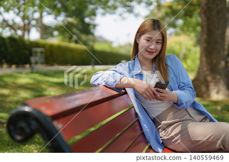 Asian woman in 20s relaxing on park bench using smartphone Asian woman in 20s relaxing on park bench using smartphone 140559469