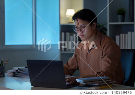 Asian man working remotely on laptop at home office at night, focused Asian man working remotely on laptop at home office at night, focused 140559476