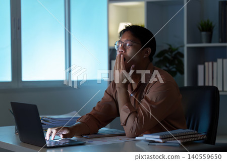 Tired Asian man yawning while working on laptop at remote home office at night 140559650